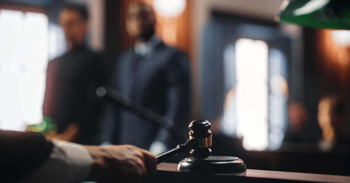 Close Up Of A Judge Banging His Gavel While A Defendant In A Suit And His Lawyer Tie Stand In The Background, Blurred And Out Of Focus