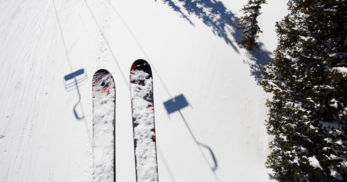 The View From A Chair Lift 25 Feet Above A Sunny, Snow-Covered Slope: A Pair of Skis, Pine Trees, And The Shadow Of The Cable And Chairs