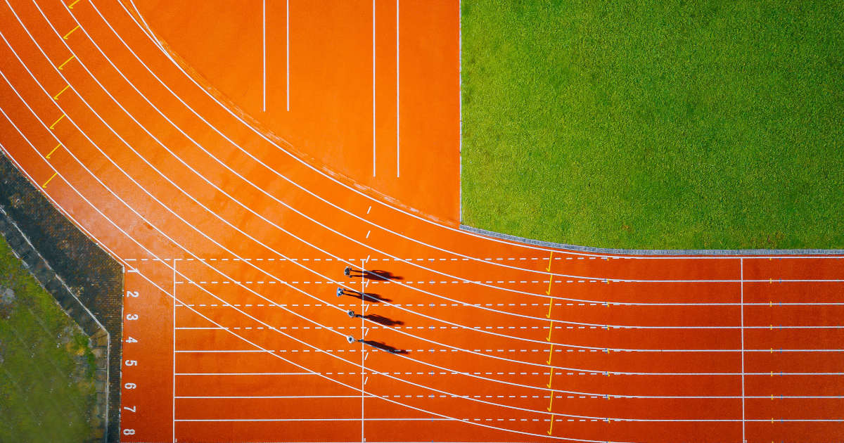 An Athletic Field Seen From Directly Overhead, The Curve of the Orange Cinder Track In The Lower Left Corner And The Green Of The Football Field In The Upper Right Corner, Four Sprinters, Tiny As Ants, Standing Near the Starting Line