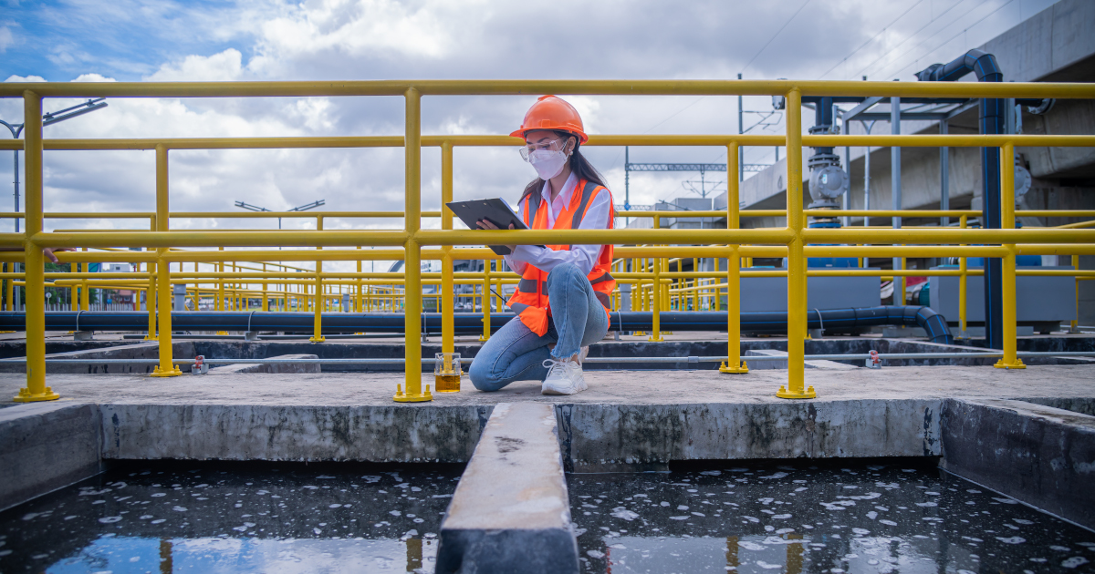 Beneath A Blue Spring Sky, A Young Woman In An Orange Hardhat And An Orange High Visibility Vest Kneels On A Concrete Walkway That Runs Through A  Wastewater Treatment Plant