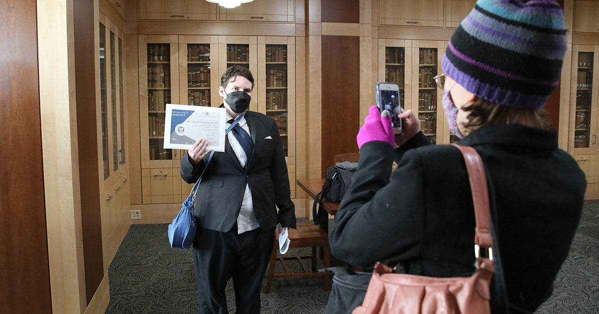 a person in a mask holds up a certificate as another person takes a photo