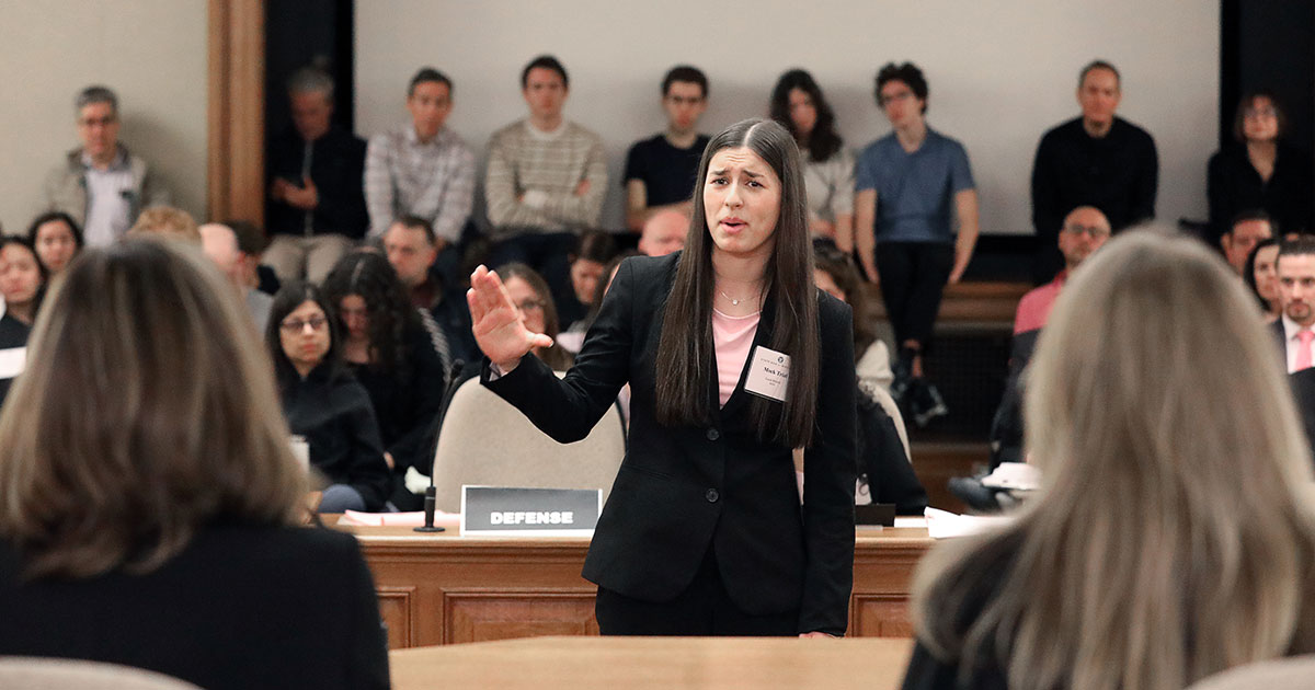 a high school student stands with hand in the air before judges with a crowd of people behind her