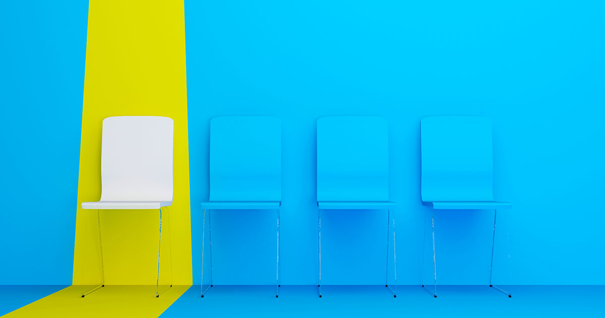Photograph of four chairs against a blue wall with a vertical yellow stripe extending onto the floor. One white chair contrasts with three blue chairs, highlighting a theme of individuality or standing out.