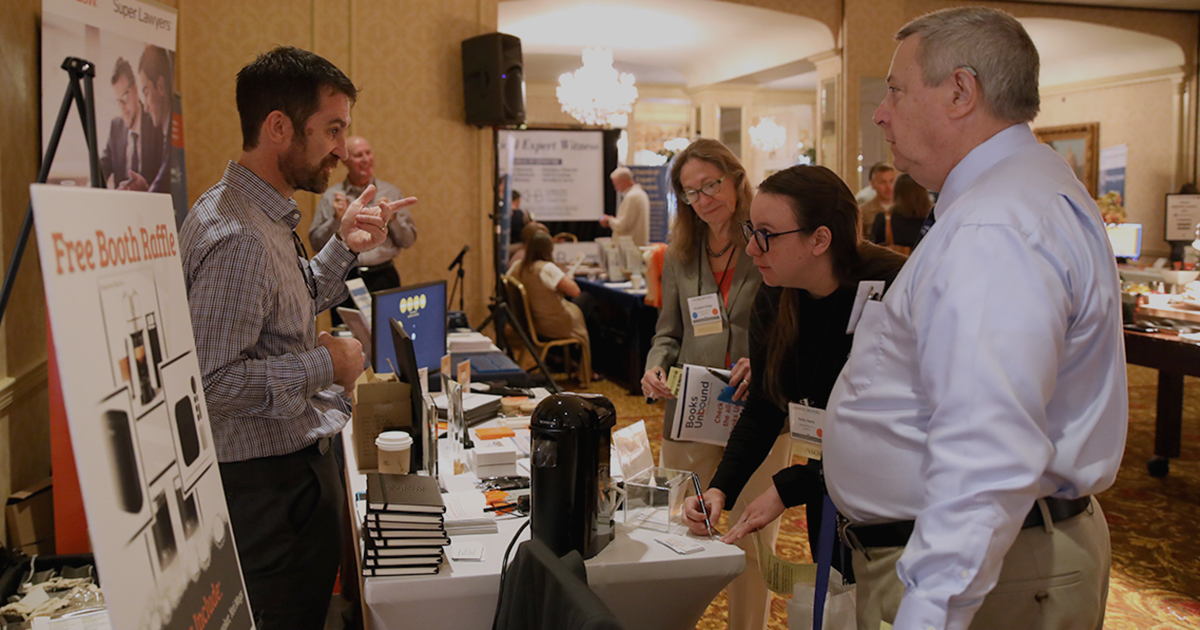 three people stand in front of a vendor's table talking with a person standing behind the table