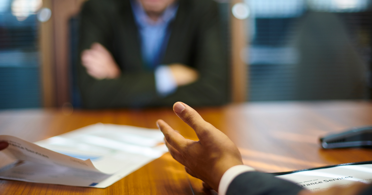 Two Men In Dark Suits Square Off Across A Honey-Colored Wooden Table, With Several Pieces of Paper Between Them. In The Foreground, A Left Arm Opening Its Palm. In the Background, Moderately Out of Focus, The Man Acorss the Table Sits With His Arms Folded.