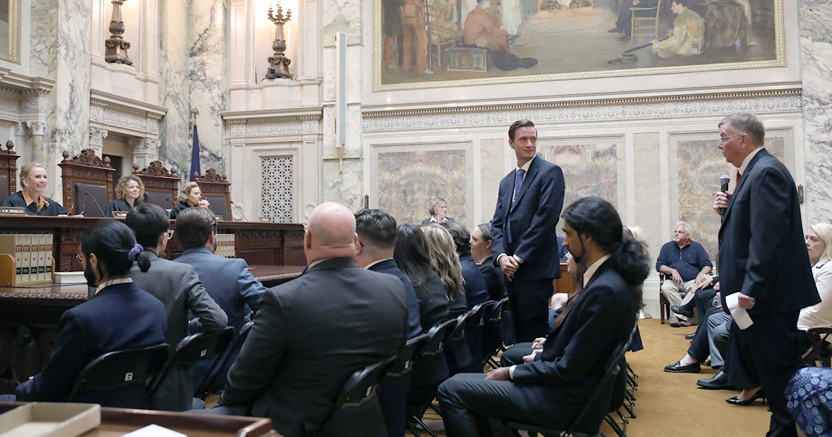 Two people are standing, one looking at the other who is holding a microphone, in the crowded Supreme Court Hearing Room