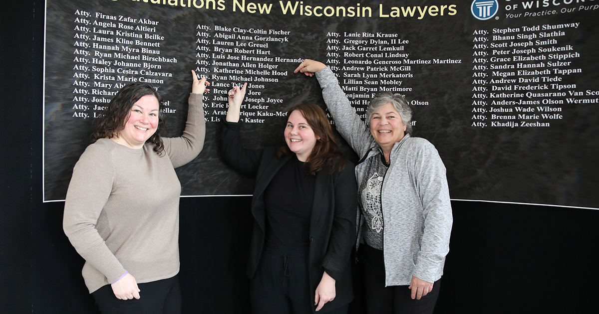 Three smiling women point to a name on a list of names behind them