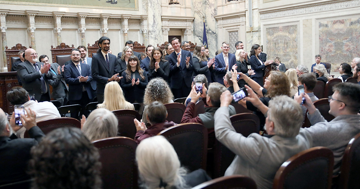 a smiling group of people are standing and clapping while facing a seated audience holding up cell phones