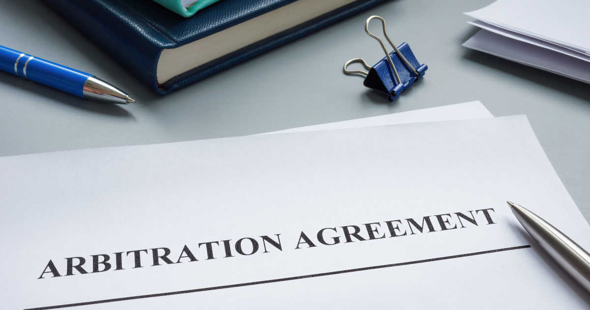 A Document Titled Arbitration Agreement Lying On A Table Top Next To Binders, Two Ball Point Pens, And A Document Clasp