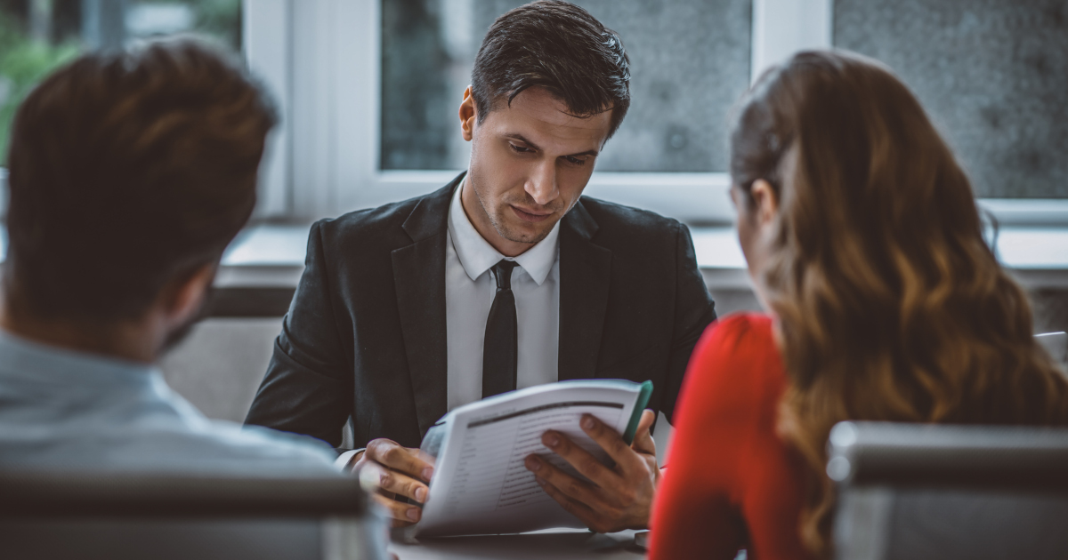 Man In Suit And Tie Reads A Document While Two People Across The Table Look On