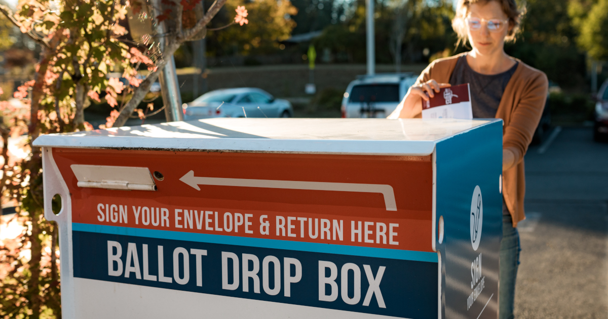 A Woman Placing A Ballot In A Ballot Drop Box, Lit By Autumn Sunlight