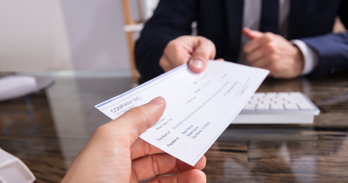 Seen Up Close, A Male Banker Wearnig A Navy Suit Coat Hands A Check Across His Desk To A Customer