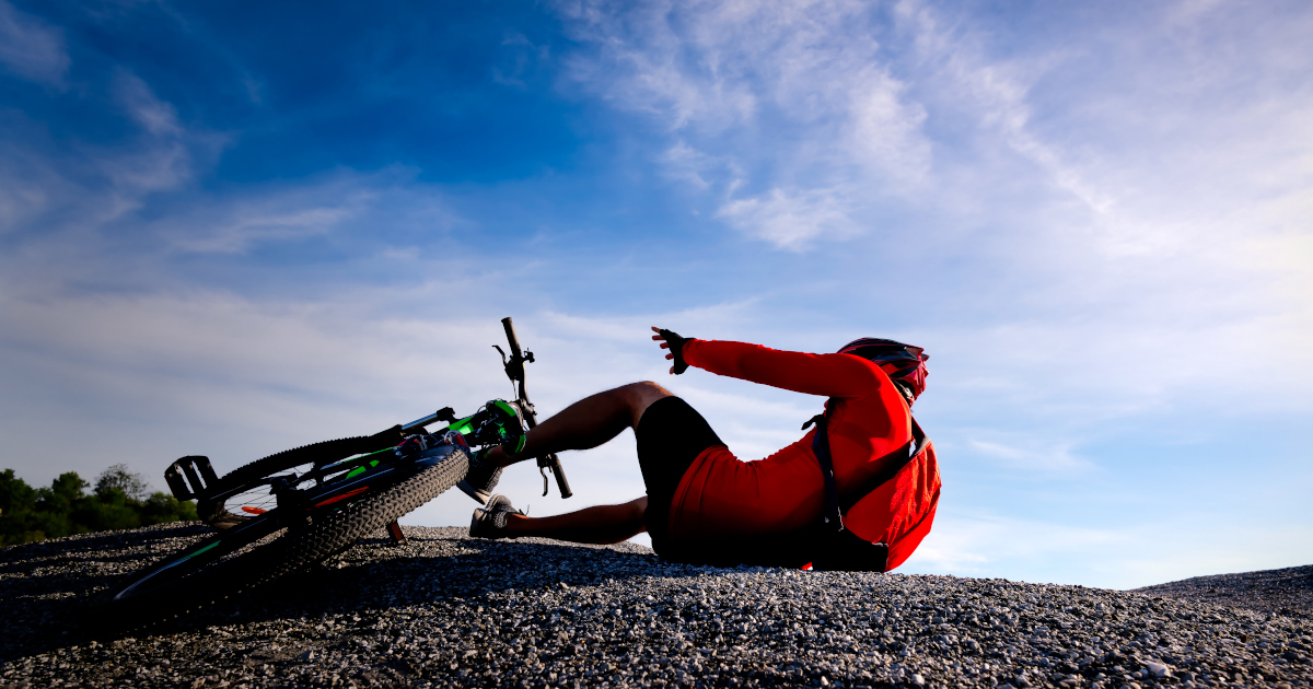 Seen From Ground Level And Against A Blue Sky, A Red-Jerseyed Bicyclist Lies Awkwardly On The Aspahlt And Reaches Out In Pain Next To His Fallen Mountain Bike