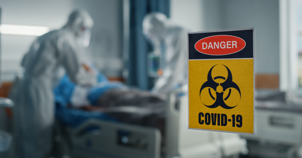 Two Nurses In Hazmat Suits Bend Over A Patient In A Hospital Bed Behind A Window Marked With A Yellow Sign That Reads Danger Covid-19