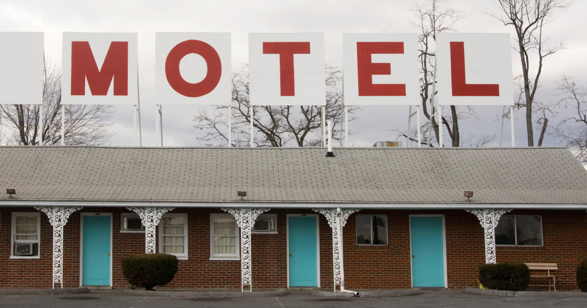 A Rundown Single Story Brick Building, With The Word Motel Spelled Out In Red Letters On A Sign Above The Shingle Roof