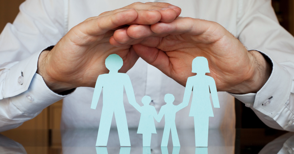 A Pair of Man’s Hands Folded Over Paper Cutouts Of A Man And Woman Holding Hands With Two Children Between Them