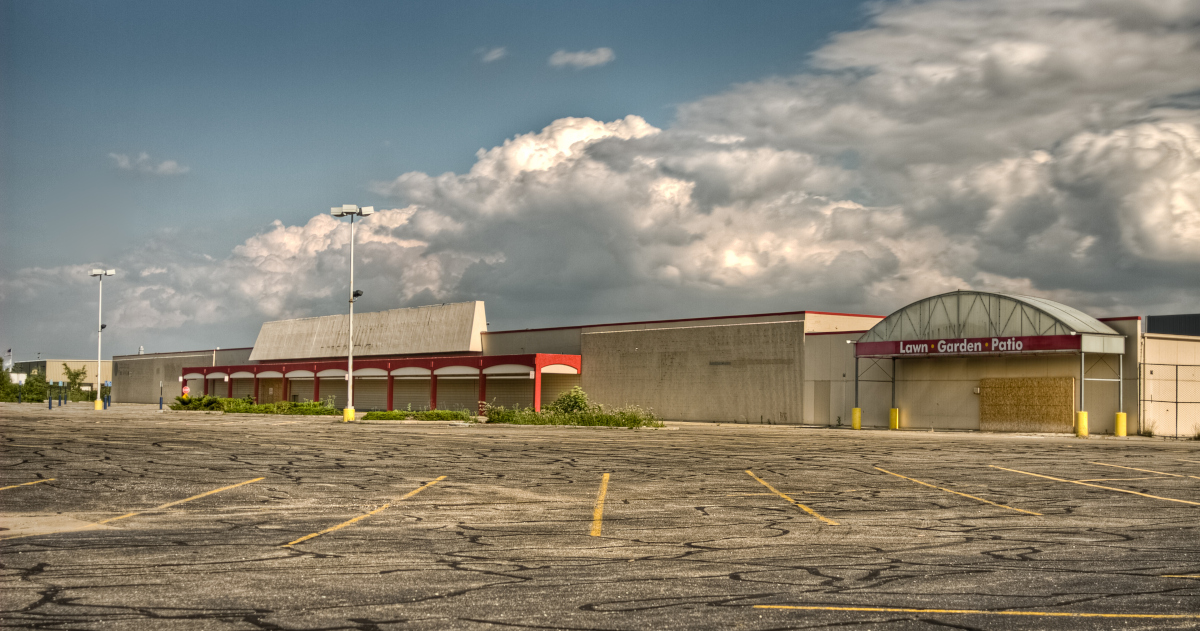 Beneath Summer Clouds, A Large Retail Store Stands Vacant Beyond A Weed-Choked Parking Lot