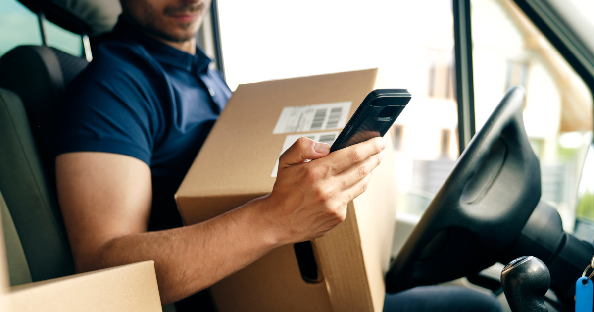 A Man In A Dark Blue Polo Shirt, Sitting In A Car Cradling a Brown Cardboard Box And Looking At His Smartphone