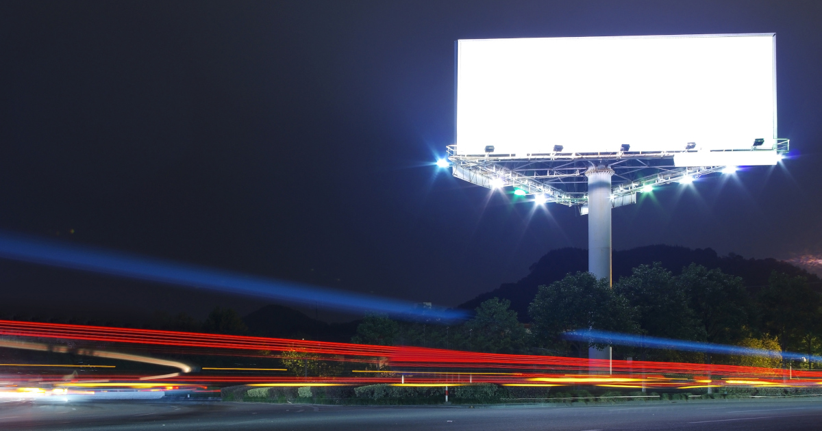 A Large Digital Billboard, Glowing White And Empty, Profiled Against The Night Sky As Taillights and Headlights Streak By Below