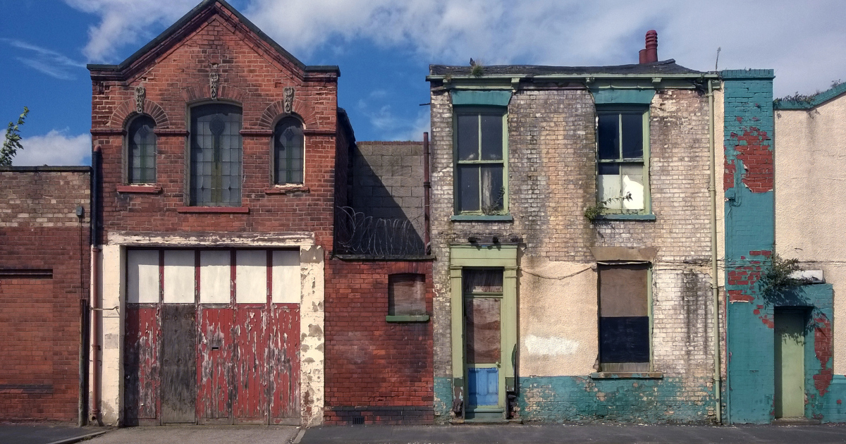 Two Brick Buildings With Flaking Paint And Broken Windows Stand Adjacent Along A City Street