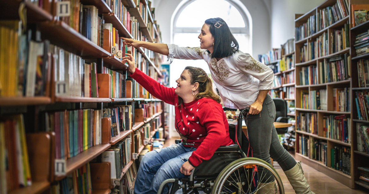 A Young Woman In A Wheelchair Reaching Toward A Book On A Library Shelf While A Young Woman Standing Behind The Wheelchair Reaches To Help Her