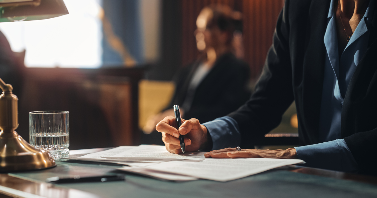 A Woman In A Navy Blue Business Suit Signing A Document On A Table In A Side-lit Courtroom