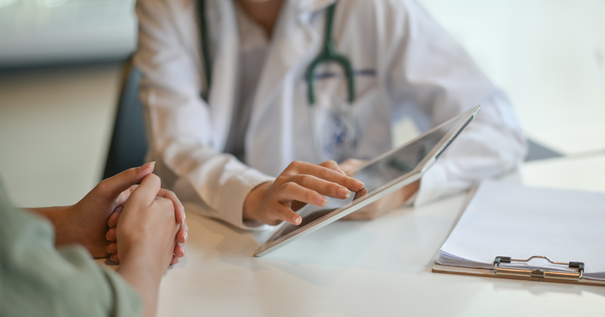 Close Up Of A Woman's Hands And Across The Table A White-Coated Woman Doctor's Hands Touching An Electronic Tablet