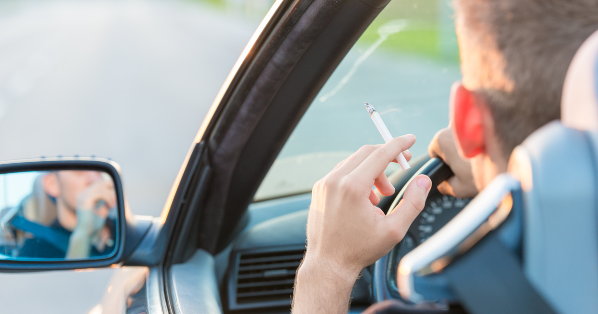 Seen From Behind And To The Left, A Man Seated In The Driver's Seat Of A Car, With The Window Open And A Freshly Lit Cigarette In His Left Hand