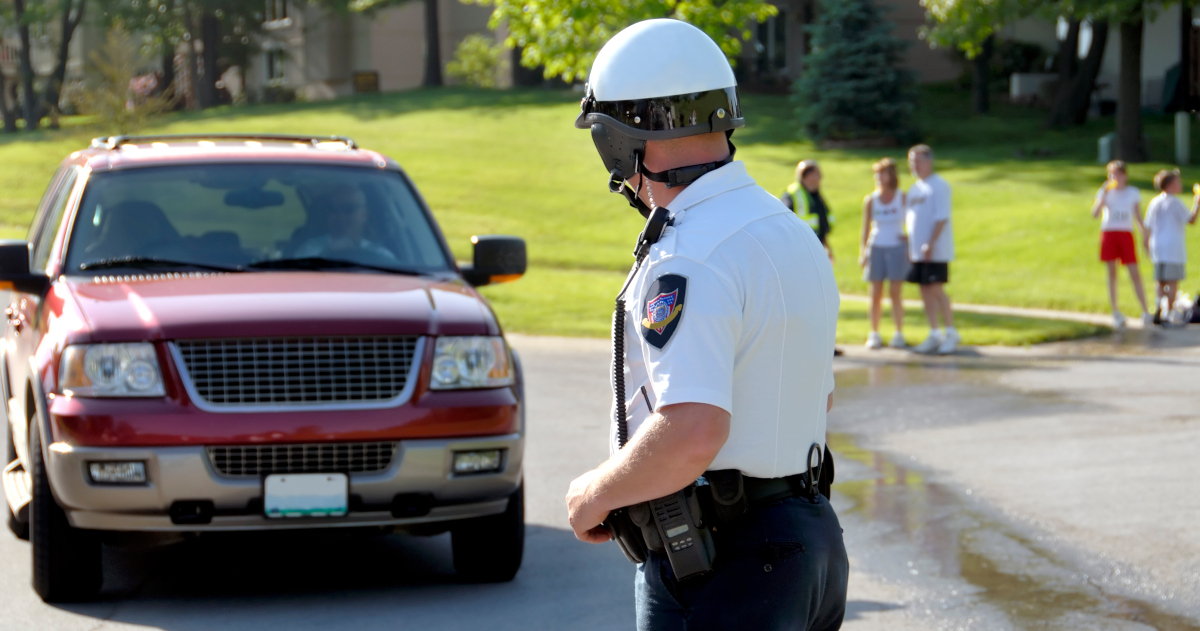 A Motorcycle Police Officer Standing In A Driveway Looking At An SUV As It Pulls In