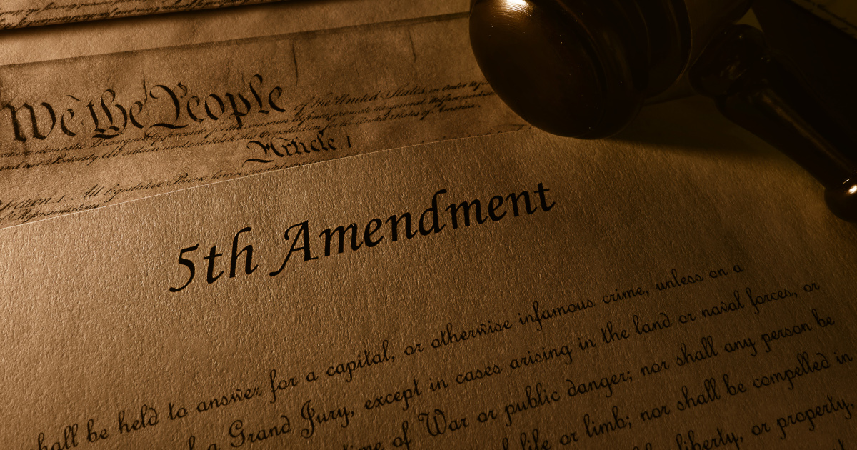 A Closeup Of A Copy Of The Declaration of Idependence And A Copy Of The Fifth Amendment, Lit by Sombre Light, With A Judge's Gavel Resting At An Angle Across Them