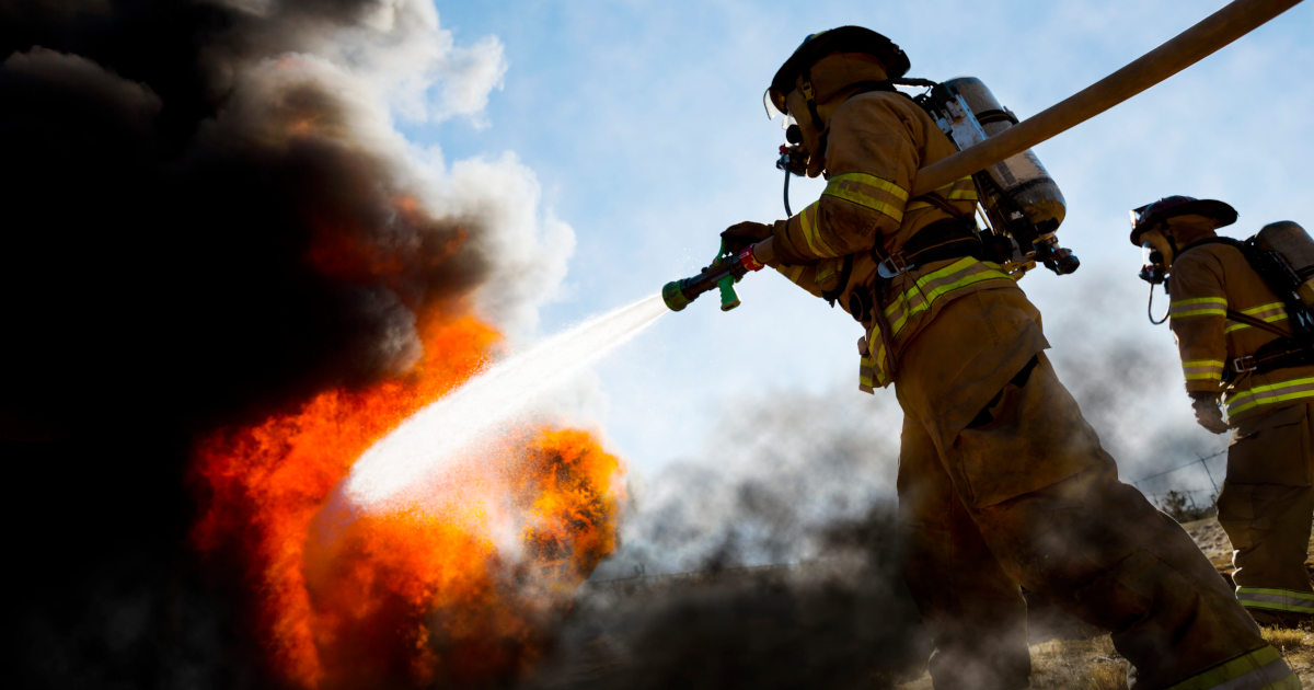 Two Firefighters In Fire Coats Battling A Blaze With A Fire Hose