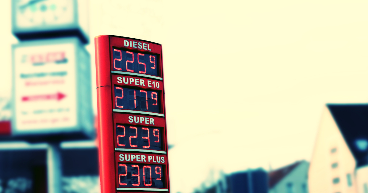 A Vertical Red Signs Displaying Prices for Diesel, Super E10, Super, Super Plus, and Autogas LP Rises Against A Backdrop Of A Convenience Store Sign and Houses In The Distance