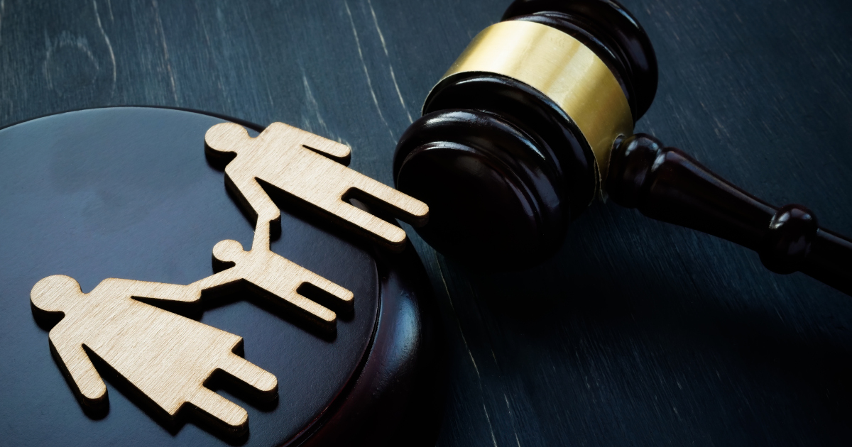 A Wooden Cutout Of A Mother And A Father Holding Hands With A Child Between Them, With A Judge's Gavel Lying Next To Them