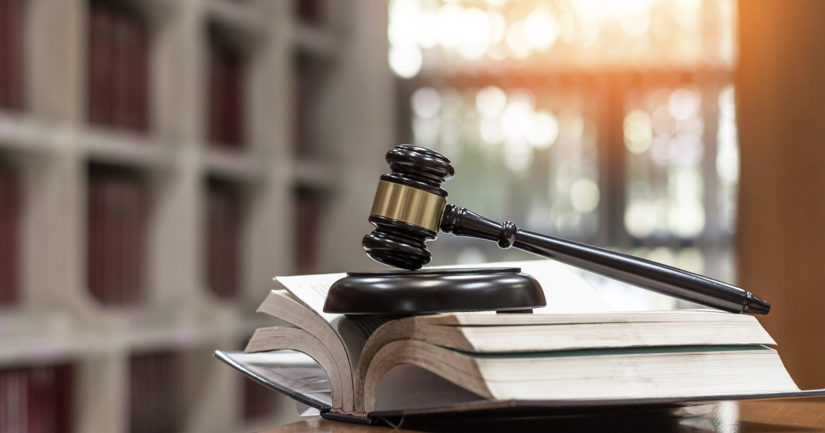 A Gavel, Resting On Its Sound Block, Balanced Atop And Propping Open A Thick Binder of Legal Forms, Backlit by Late Afternoon Light