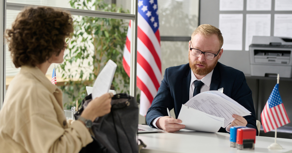 A Woman Standing In Front Of A Counter, Behind Which Sits A Man Reviewing A Stack Of Papers, With An American Flag On A Pole Behind Him