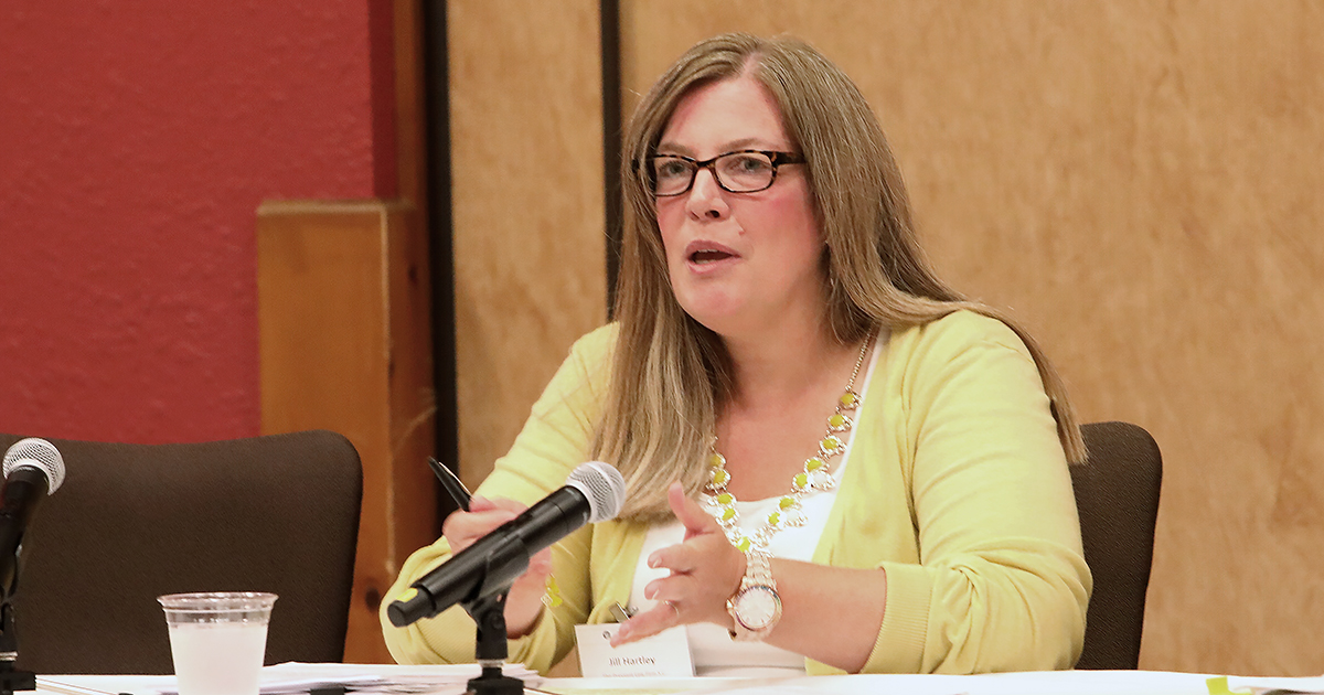 A Woman Seated Behind A Table Speaking Into A Tabletop Microphone