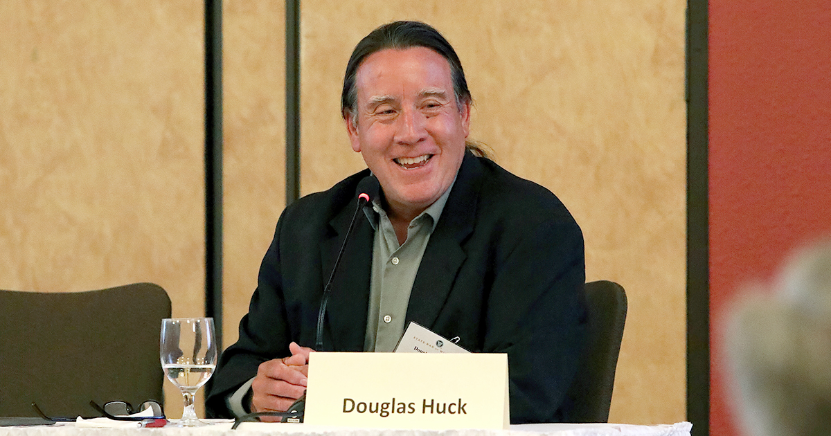 A Dark-Haired Man With A Ponytail Speaks Into A Tabletop Microphone While Gesturing With One Hand