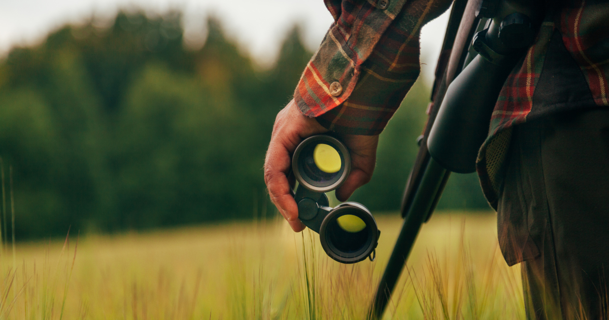 Closeup Of A Man In A Flannel Shirt Holding in His Left Hand A Pair of Binoculars With A Rifle Slung Over His Shoulder, While Standing In A Field of Hip-High Grass