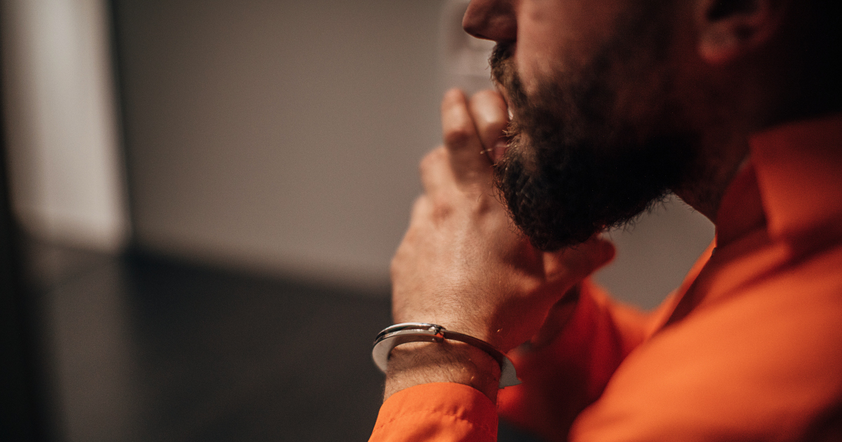 A Bearded Male Prisoner In An Orange Jumpsuit and Handcuffs Sits In A Prison Cell