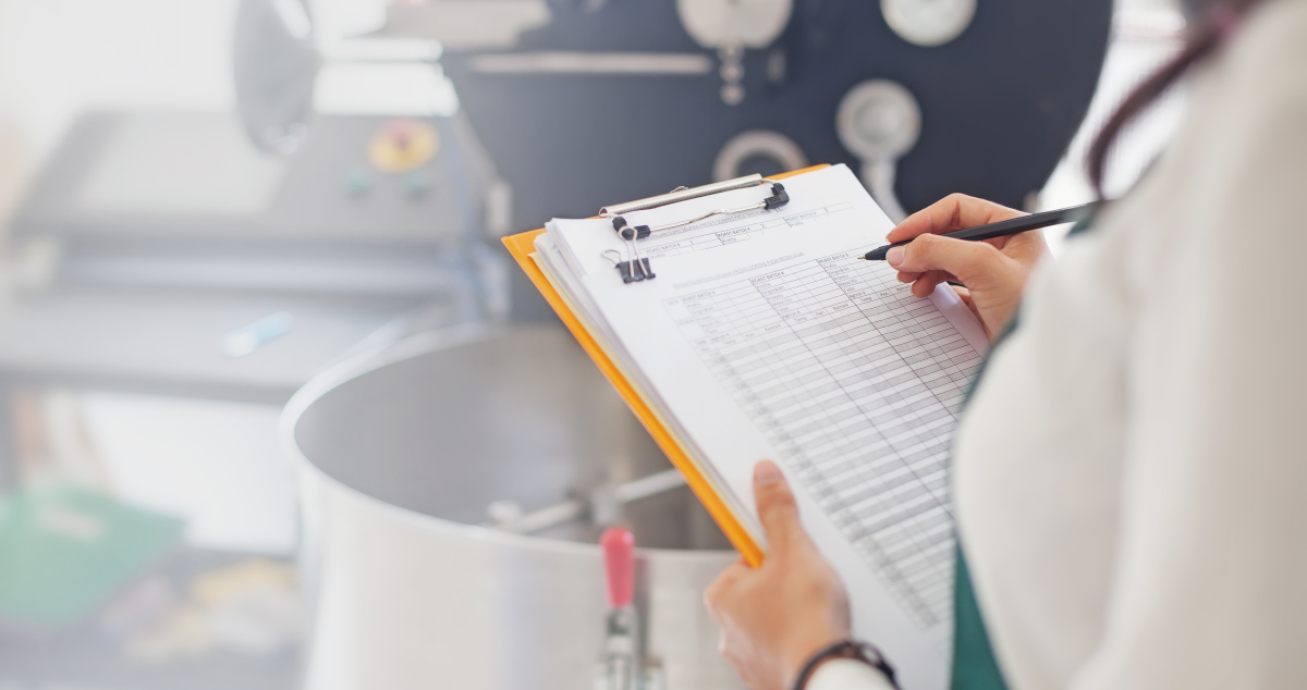 A Woman In A White Lab Coat Holding A Clipboard And Checklist in One Hand And A Pen In The Other, As She Stands In Front Of A Steaming Vat With Pressure Gauges