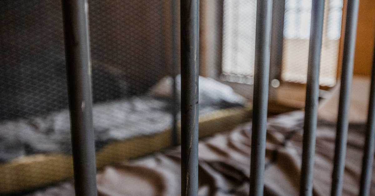 Two Beds In A Jail Cell, Lit By Winter Light Bleeding In Through A Chicken-Wire Window