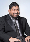 Hamza Jaka, a Brown disabled man wearing a suit and tie, sits in his wheelchair and smiles