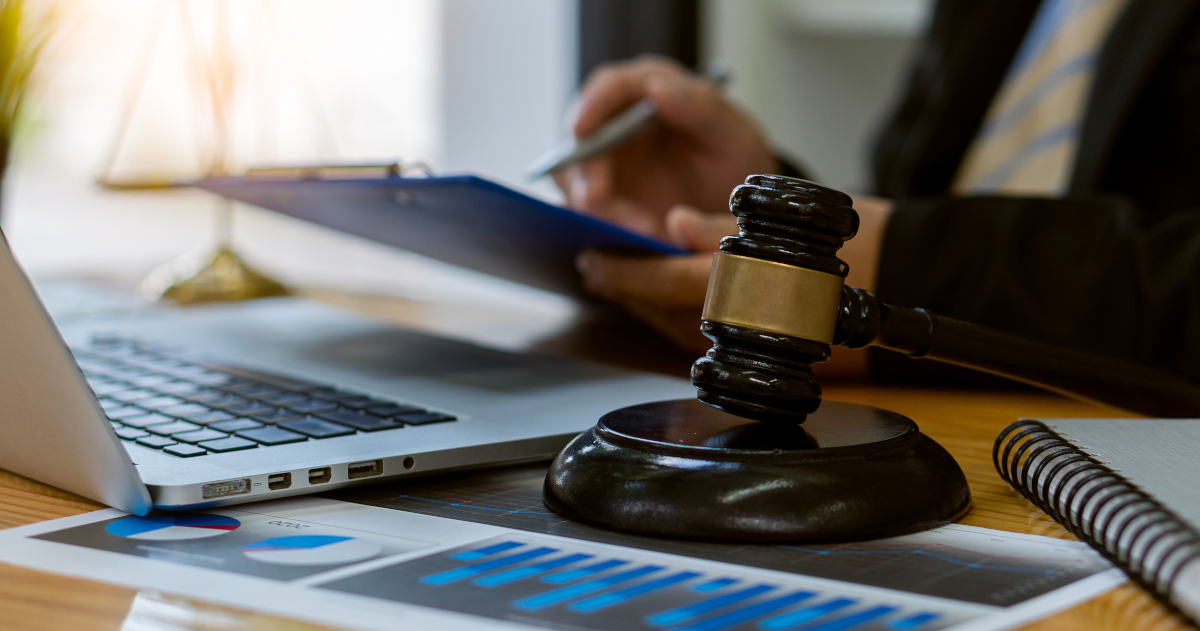 A Judge Sits In His Daylit Chambers, A Pen Poised Above A Clipboard And A Scales of Justice On His Right And A Laptop On His Right On The Table In Front Of Him