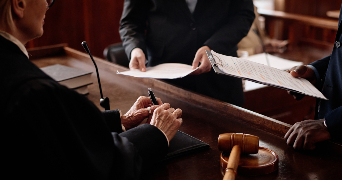 Close Up Made From Over A Judge's Right Shoulder, Showing A Woman Judge In A Black Robe Gripping A Pen In Her Hands With Her Gavel At Her Right, And Two Lawyers Stadning In Front Of The Bench, Each Holding a Sheaf of Papers