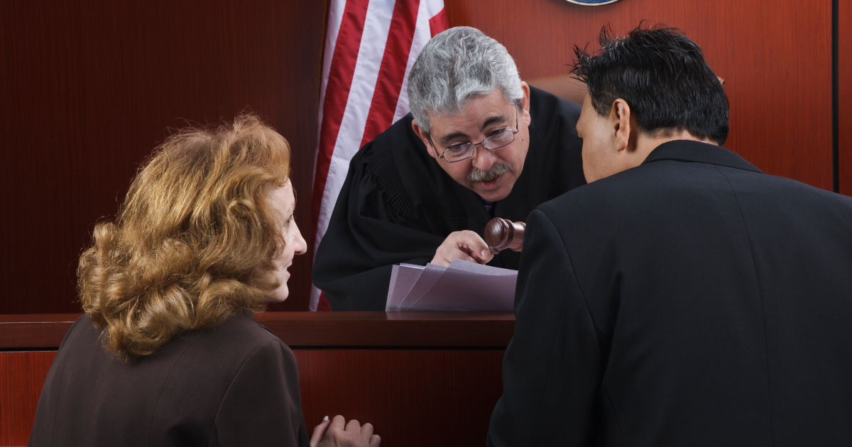 Two Lawyers, Standing Close Together With Their Heads Bent, Argue A Point Before A Judge In Front Of His Dais