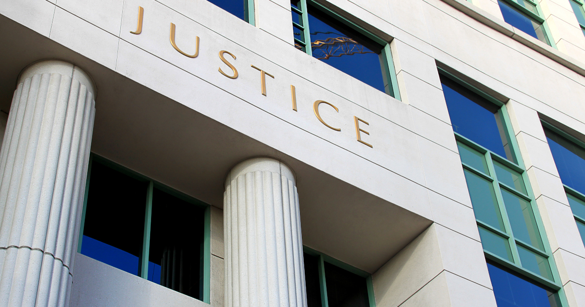 The White Stone Facade Of A Courthouse, With The Word Justice in Gold Capital Letters Above Portico Columns