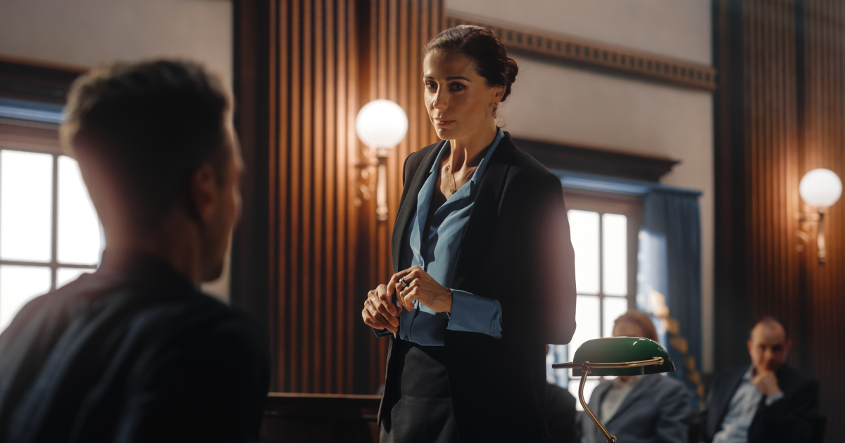 A Woman Lawyer In A Dark Gray Suit With Her Hair Pulled Back Questioning A Wintess In Court