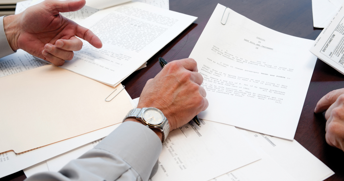 The Forearms Of Two Men In Dress Shirts, One Clutching A Pen, Reivew Legal Papers Spread Out On A Dark Wooden Desk