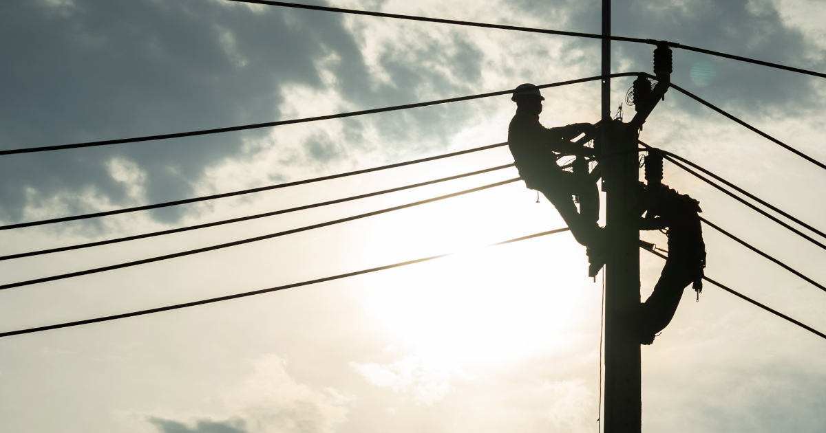 Two Electrical Linemen, Harnessed Atop A Power Pole, Silhouetted Against The Sun
