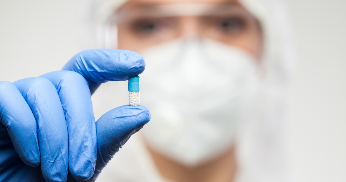 A Nurse In A Faceshield, Hood, and Surgical Mask Holding Out To The Camera a Pill Man